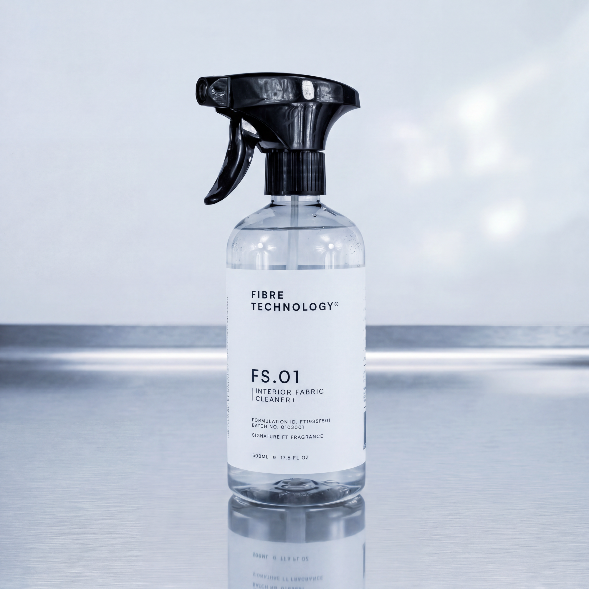 A clear spray bottle of FIBRE TECHNOLOGY FS.01 Interior Fabric Cleaner with a black trigger sprayer and white label, set on a reflective surface against a soft, out-of-focus light gray background.